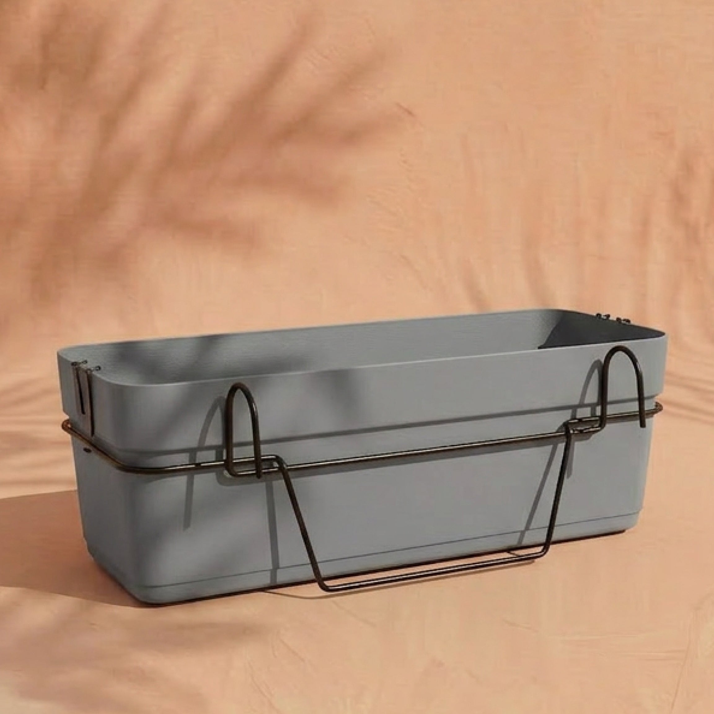 Der Blossom 60 cm Pflanzkasten für Geländer in Grau mit schwarzem Metallhalter und integriertem Wasserreservoir ruht auf einer hellen pfirsichfarbenen Oberfläche. Weiche Schatten und Blattmuster auf der pfirsichfarbenen Wand vermitteln eine gemütliche Balkonatmosphäre.