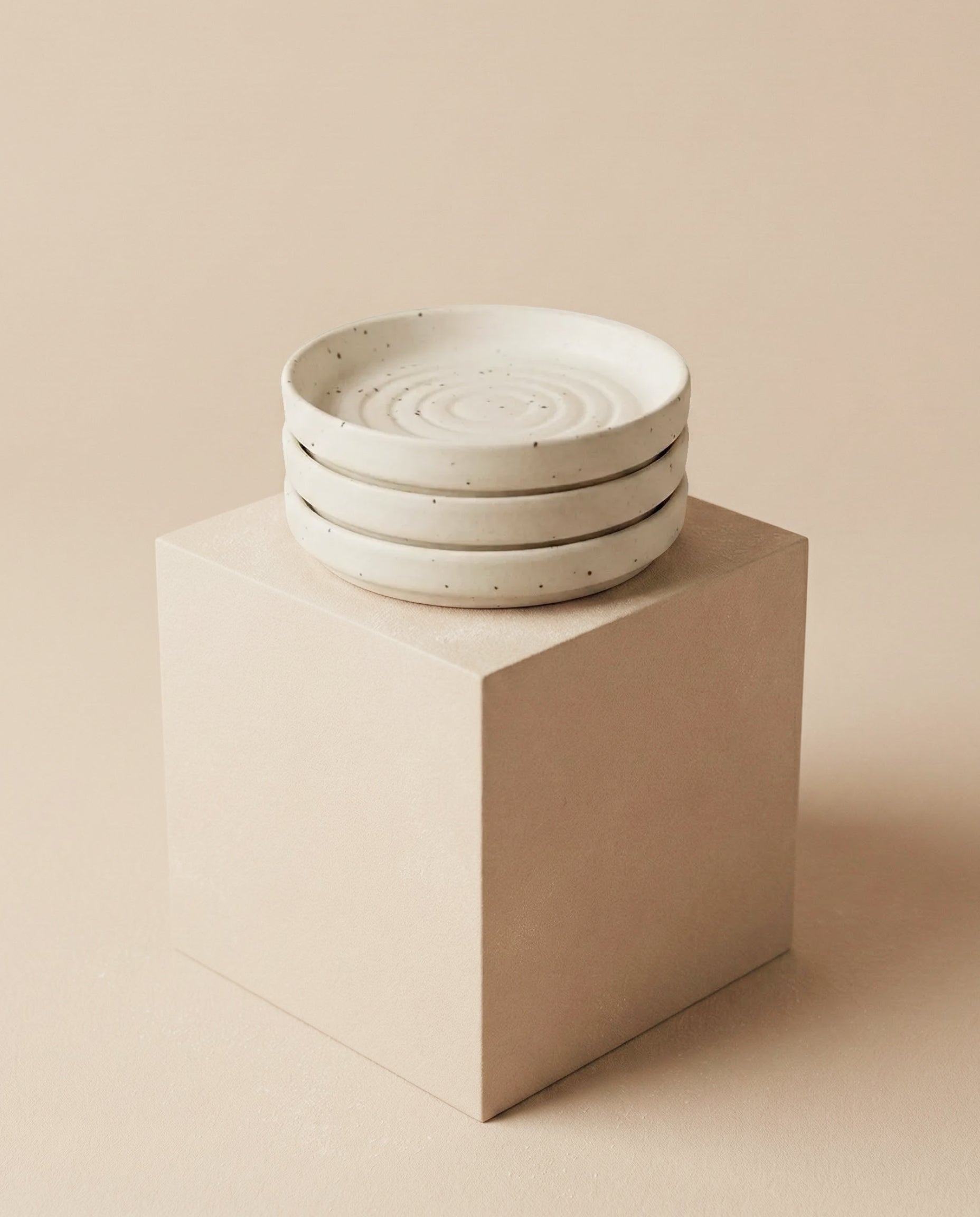 Drei Babypflanzen-Untersetzer aus Keramik (Plattino | 3 Stk.) in Weiß mit feinen Sprenkeln und kreisförmigem Muster liegen gestapelt auf einem hellbeigen Würfel vor neutralem Hintergrund.