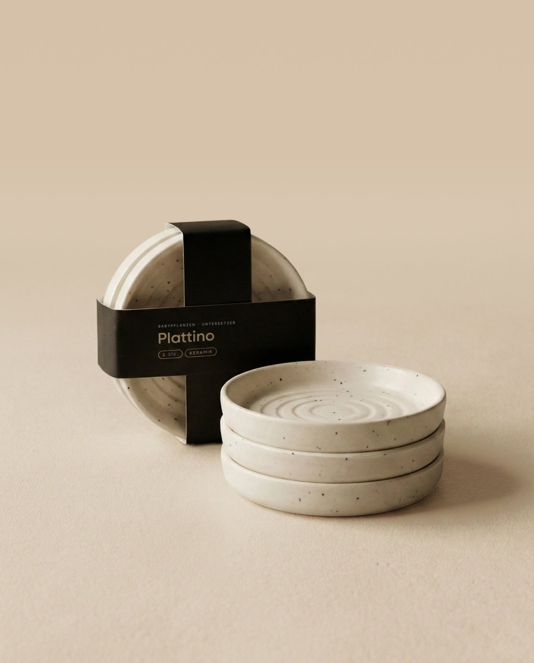 Drei gestapelte Babypflanzen-Untersetzer aus Keramik (Plattino | 3 Stk.) mit einem leicht gesprenkelten Muster sitzen neben einem passenden Set, das mit einem schwarzen 