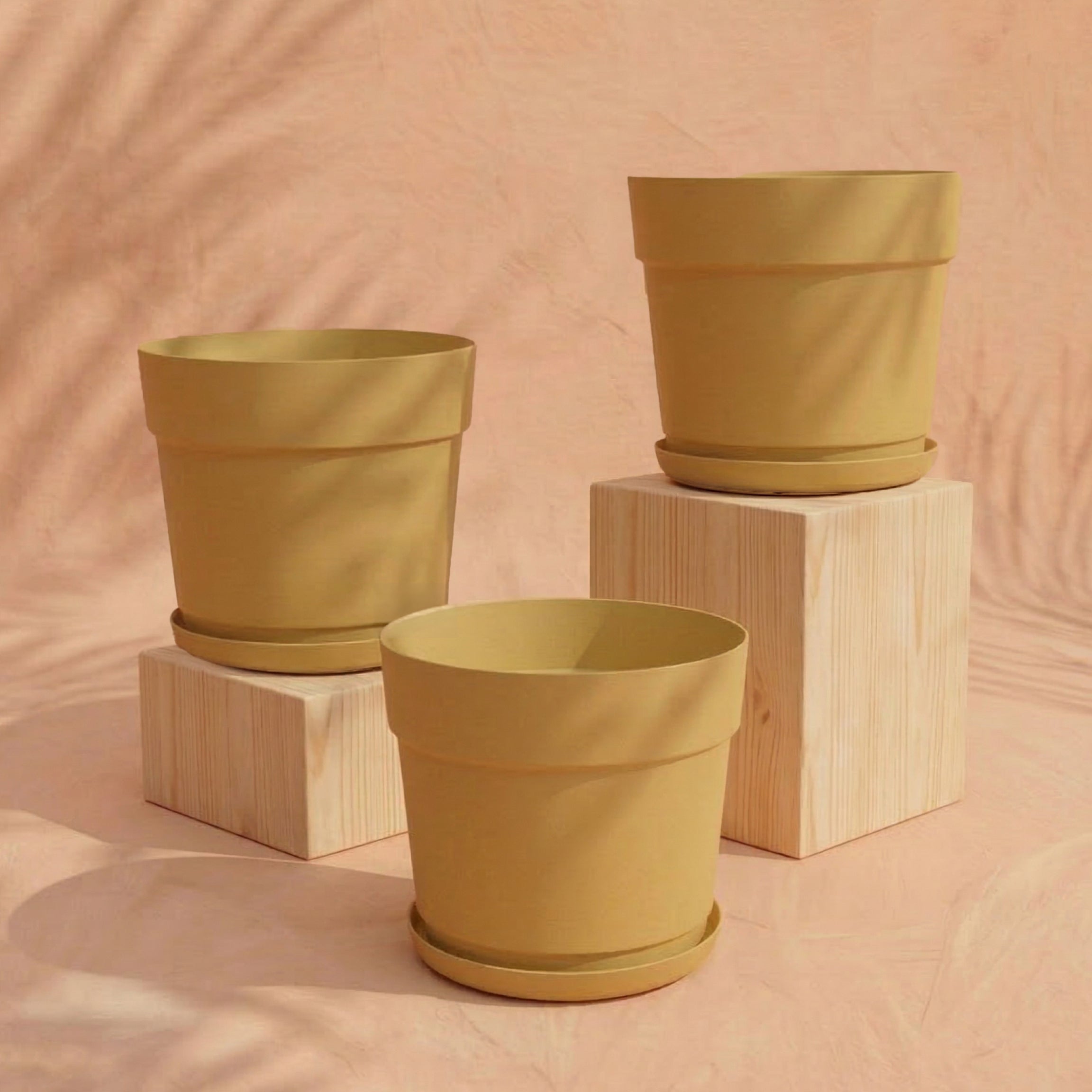 Drei gelbe Pflanzkübel aus recyceltem Kunststoff aus dem Pflanzkübel-Set 'Bloom' (3× 29) stehen auf hellen Holzblöcken vor pfirsichfarbenem Hintergrund. Palmblattschatten erzeugen eine sanfte, natürliche Atmosphäre.
