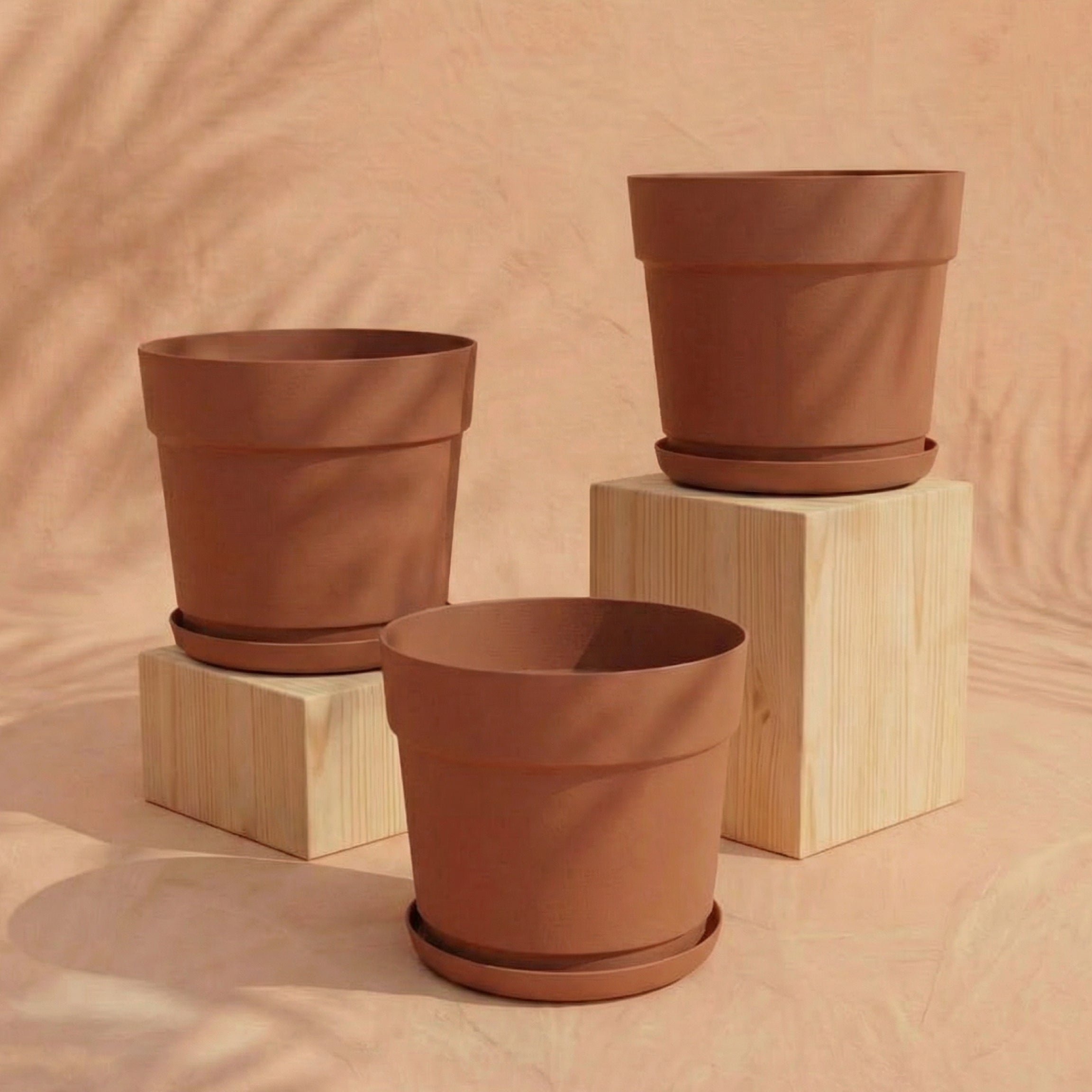 Das Pflanzkübel-Set 'Bloom' (3× 29) - drei helle, UV-beständige Terrakotta-Töpfe mit passenden Untersetzern - ist auf und neben zwei hellen Holzblöcken vor einem beigen Hintergrund mit sanften Schatten arrangiert.