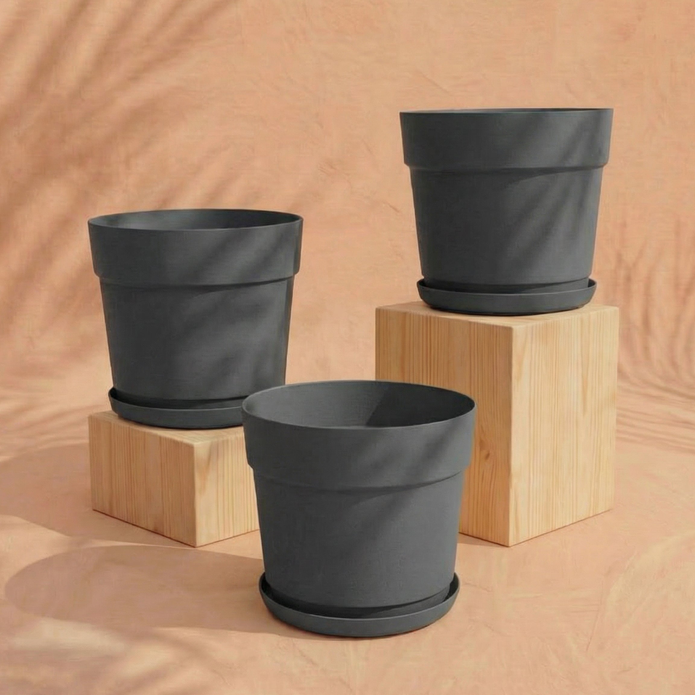 Drei leere schwarze Pflanzkübel-Sets 'Bloom' (3× 29) mit passenden Untertassen aus recyceltem Kunststoff sind ausgestellt: zwei auf hellen Holzblöcken und eine davor, alle vor einem beigen Hintergrund.