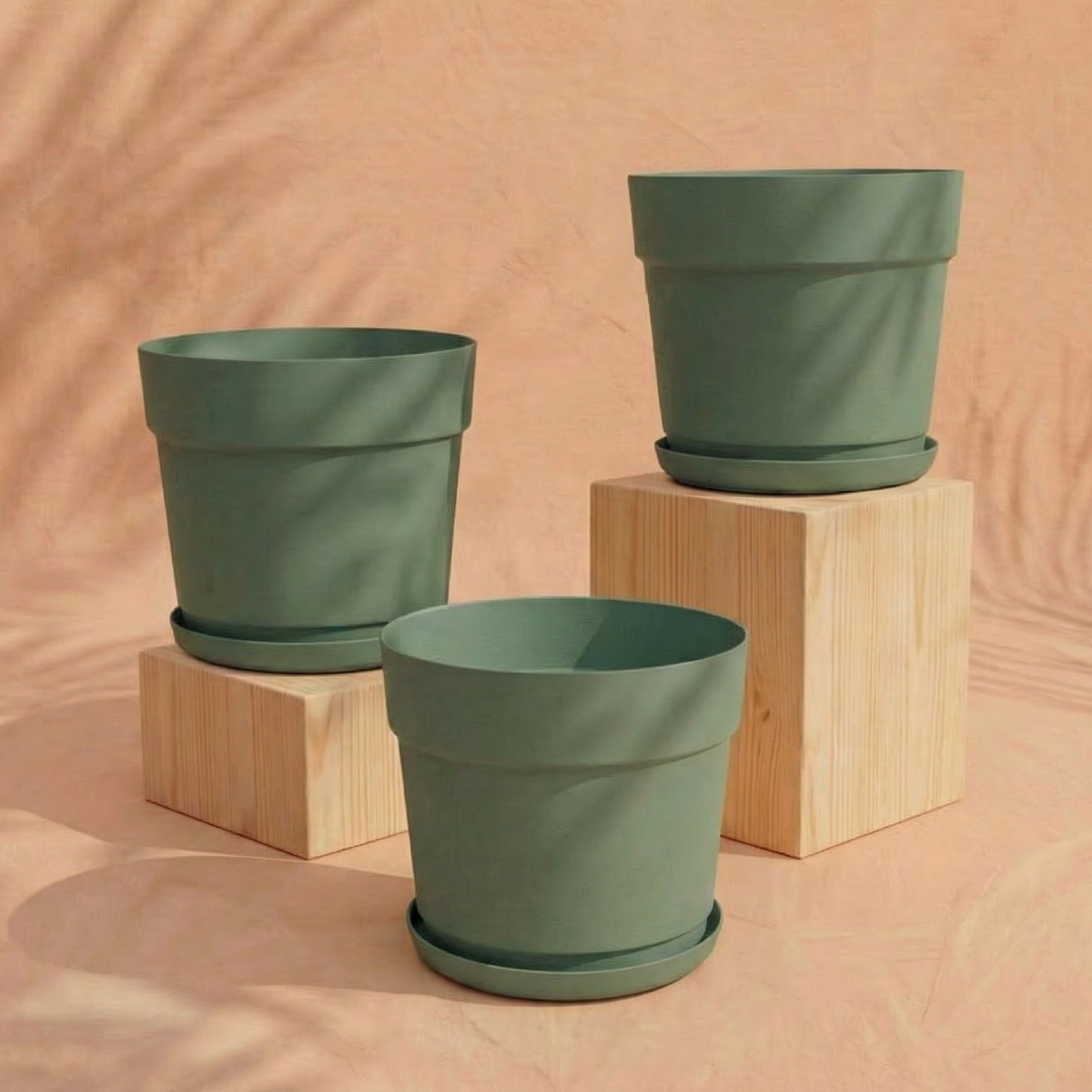 Das Pflanzkübel-Set 'Bloom' (3× 29) aus recyceltem Kunststoff mit passenden Untersetzern steht auf und neben hellen Holzblöcken vor pfirsichfarbenem Hintergrund. Die Szene ist sanft beleuchtet, UV-beständig und wirft dezente Schatten.