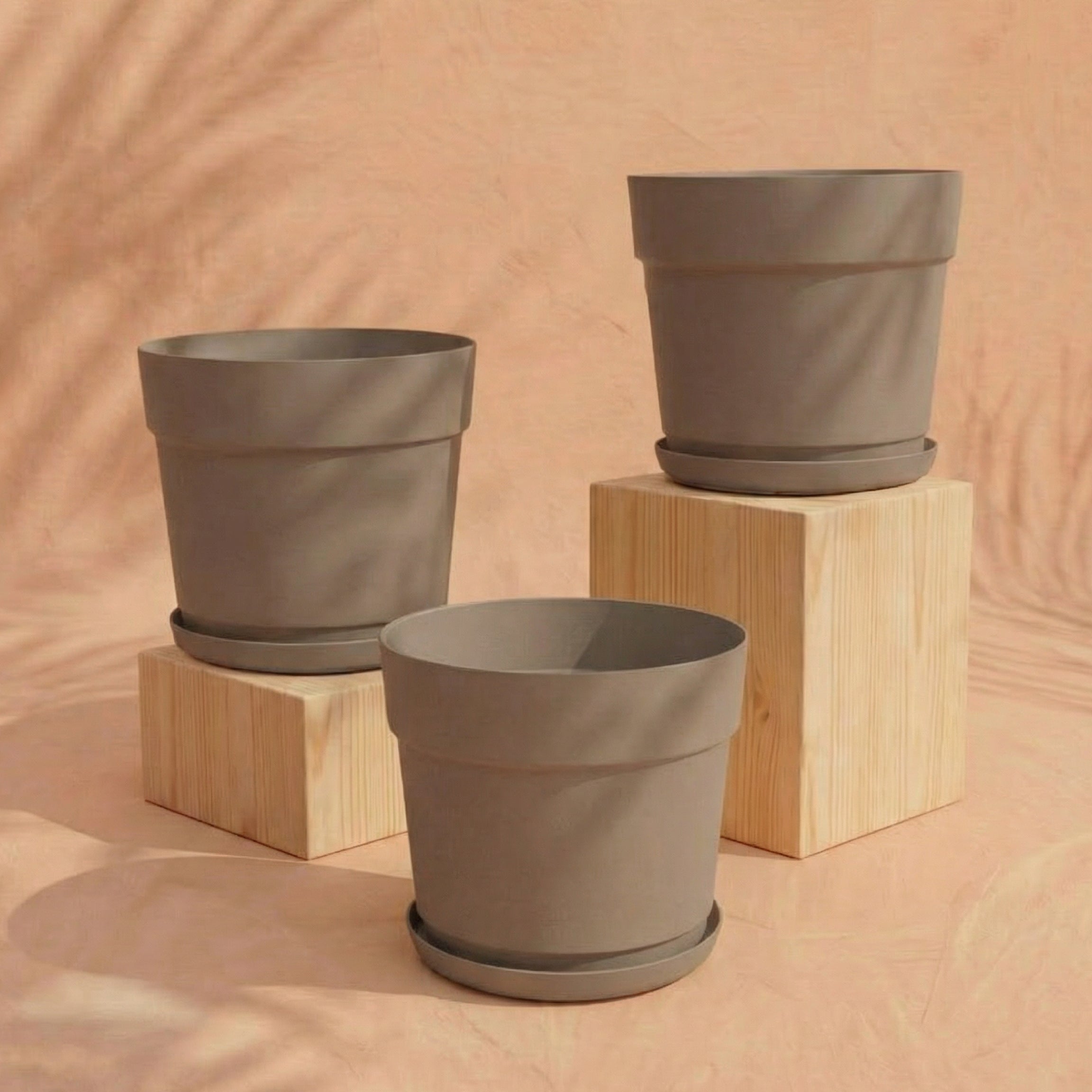 Das Pflanzkübel-Set 'Bloom' (3× 29) aus recyceltem Kunststoff mit passenden Untersetzern steht auf hellen Holzblöcken vor pfirsichfarbenem Stoff - nachhaltig und UV-beständig.