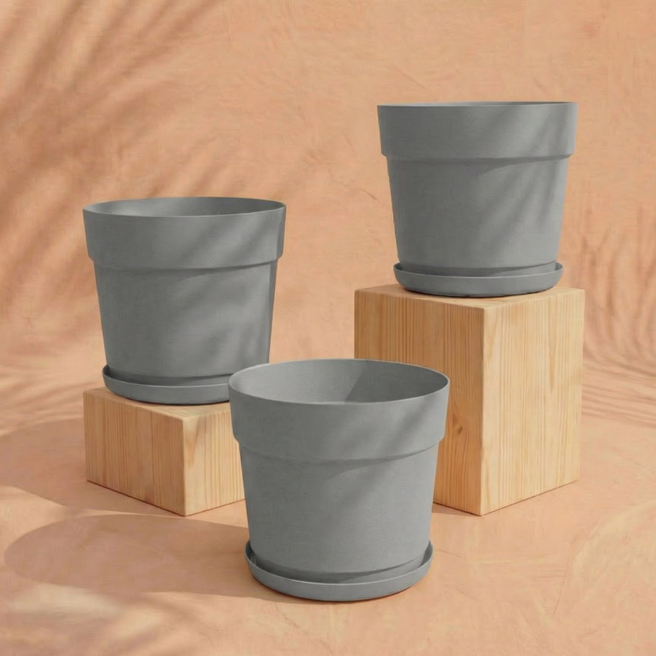 Das Pflanzkübel-Set 'Bloom' (3× 29) in Grau mit passenden Untersetzern steht auf zwei hellen Holzblöcken vor einem pfirsichfarbenen, sanft strukturierten Hintergrund mit dezenten Schattenmustern.