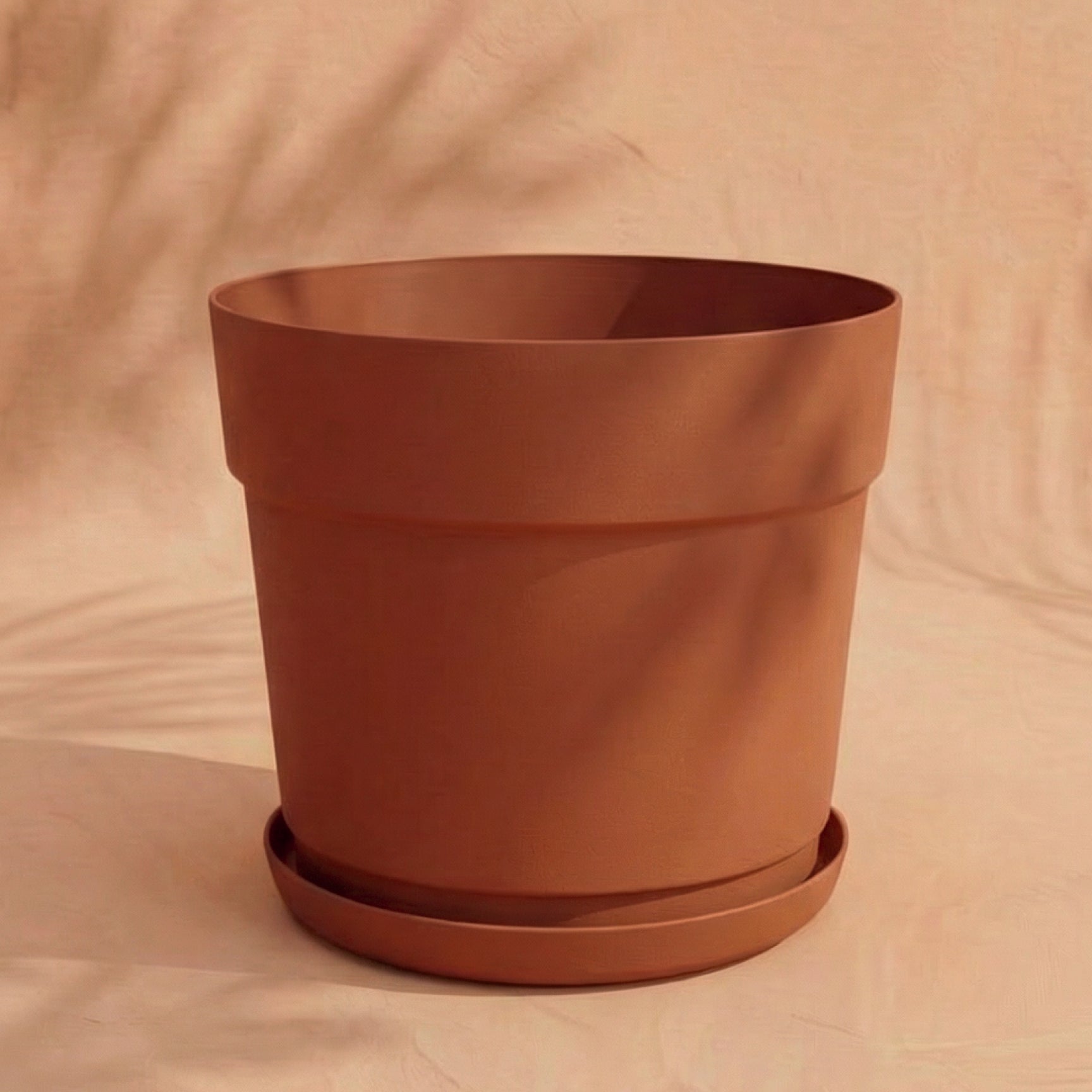 Ein großer Kunststoff Pflanzkübel (Bloom | 29 cm Ø) mit passendem Untersetzer steht auf einer neutral beigen Fläche und wirft sanfte Schatten im warmen, natürlichen Licht.