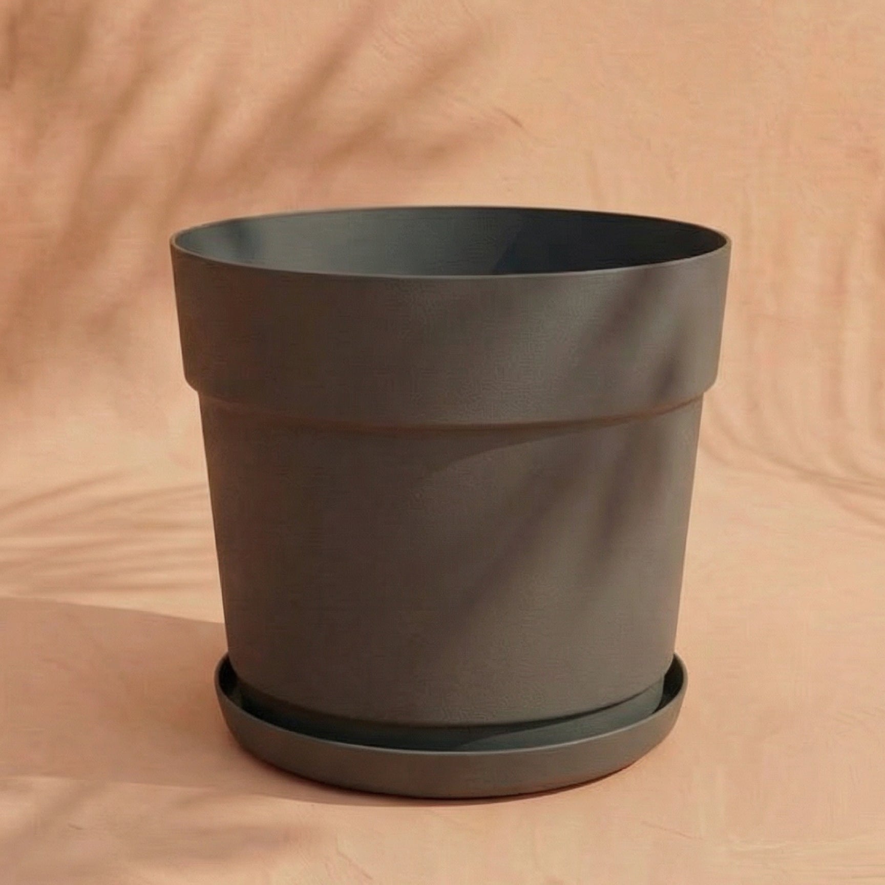 Ein großer, leerer Kunststoff Pflanzkübel (Bloom | 29 cm Ø) aus recyceltem Material mit passendem Untersetzer steht auf einer neutral beigen Fläche, weiche Schatten schaffen einen sanften Hintergrund.