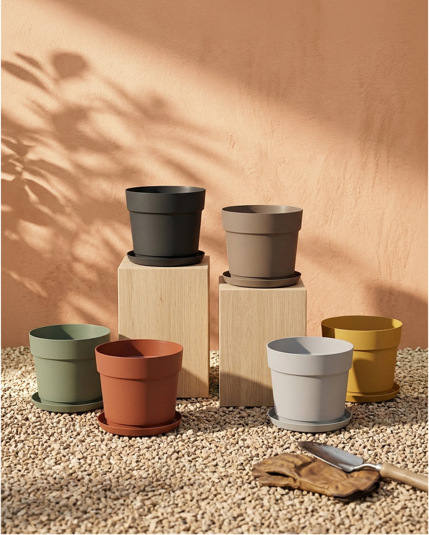 Sechs bunte Kunststoff Pflanzkübel (Bloom | 19 cm Ø) mit UV-beständiger Untertasse stehen auf Kies. Drei Töpfe sind auf Holzplinthen erhöht. Neben ihnen liegen ein Gartenhandschuh und eine Kelle, Blattschatten fällt auf die Pfirsichwand.