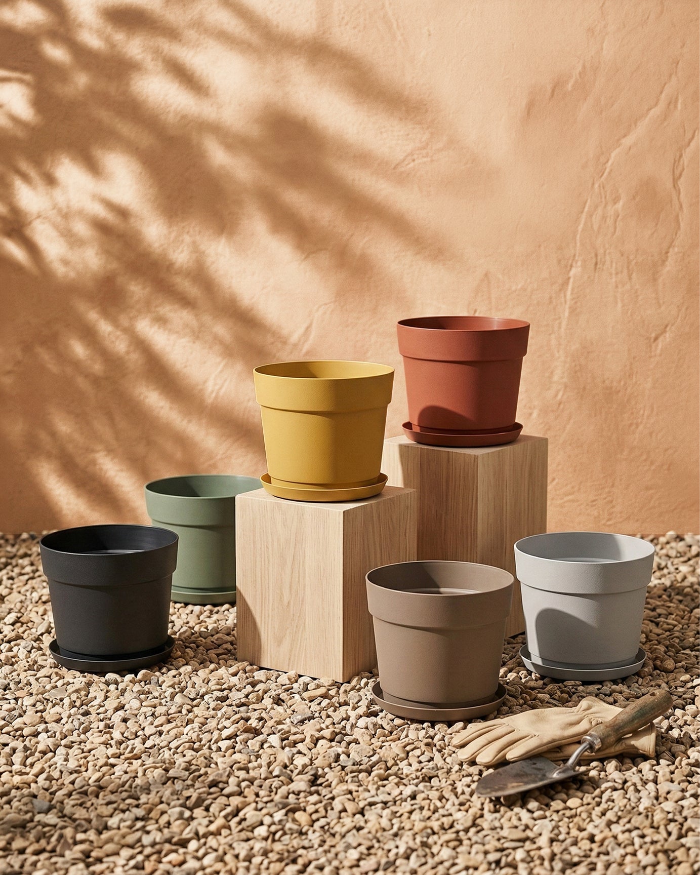 Sechs Kunststoff Pflanzkübel (Bloom | 19 cm Ø) aus recyceltem Material stehen auf Holzblöcken und Kieseln vor einer Pfirsichwand. Gartenhandschuhe und Werkzeuge liegen daneben, Sonnenlicht wirft Schatten auf die UV-beständigen Töpfe.