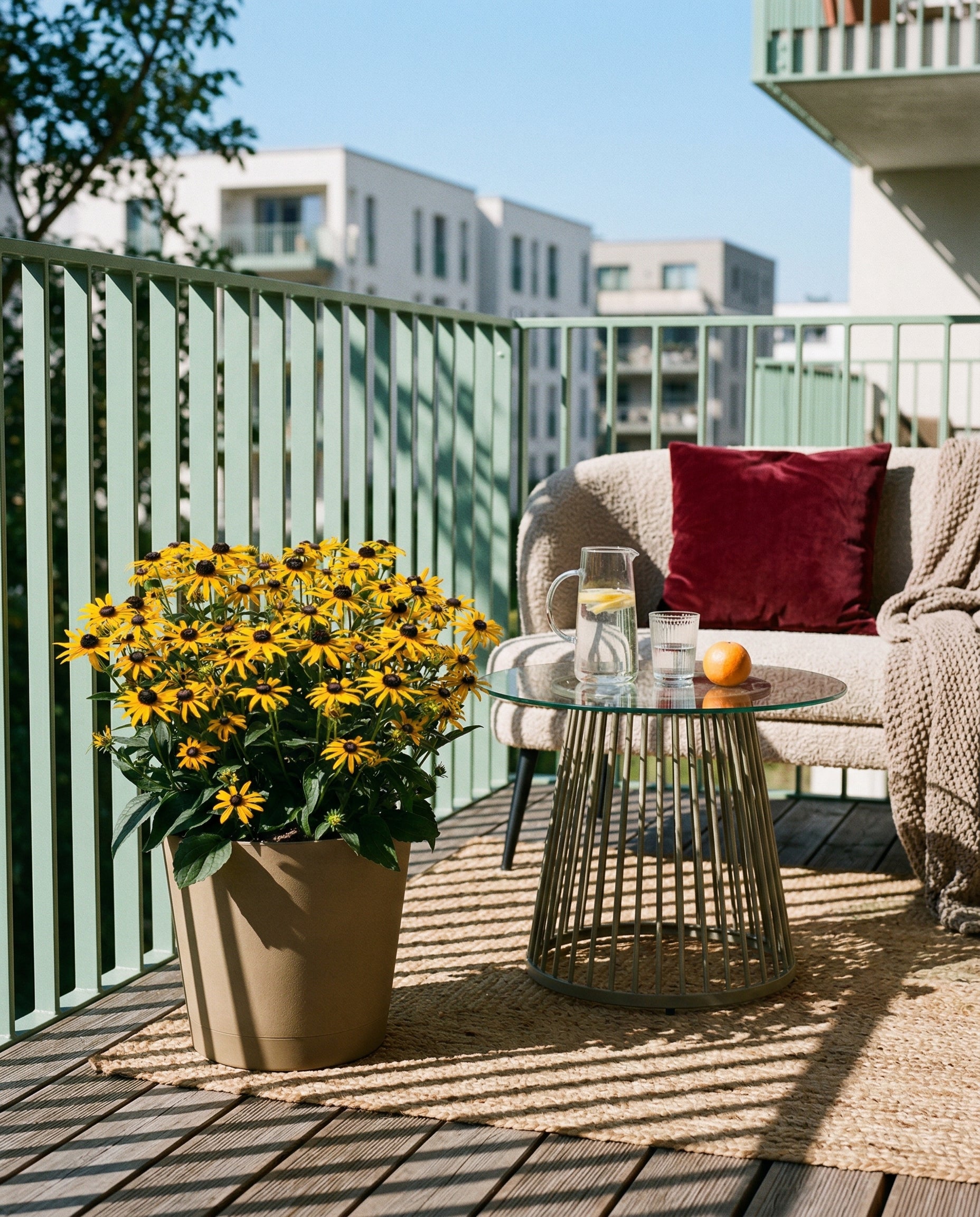 Auf dem gemütlichen Balkon stehen ein Stuhl mit rotem Kissen, eine beigefarbene Decke, ein Glastisch mit Wasser, eine Kerze, eine Orange und ein großer, getöpferter Gelber Sonnenhut (Wurzelstock), dessen gelbe Blüten den Raum vor den Gebäuden der Stadt und dem blauen Himmel aufhellen.