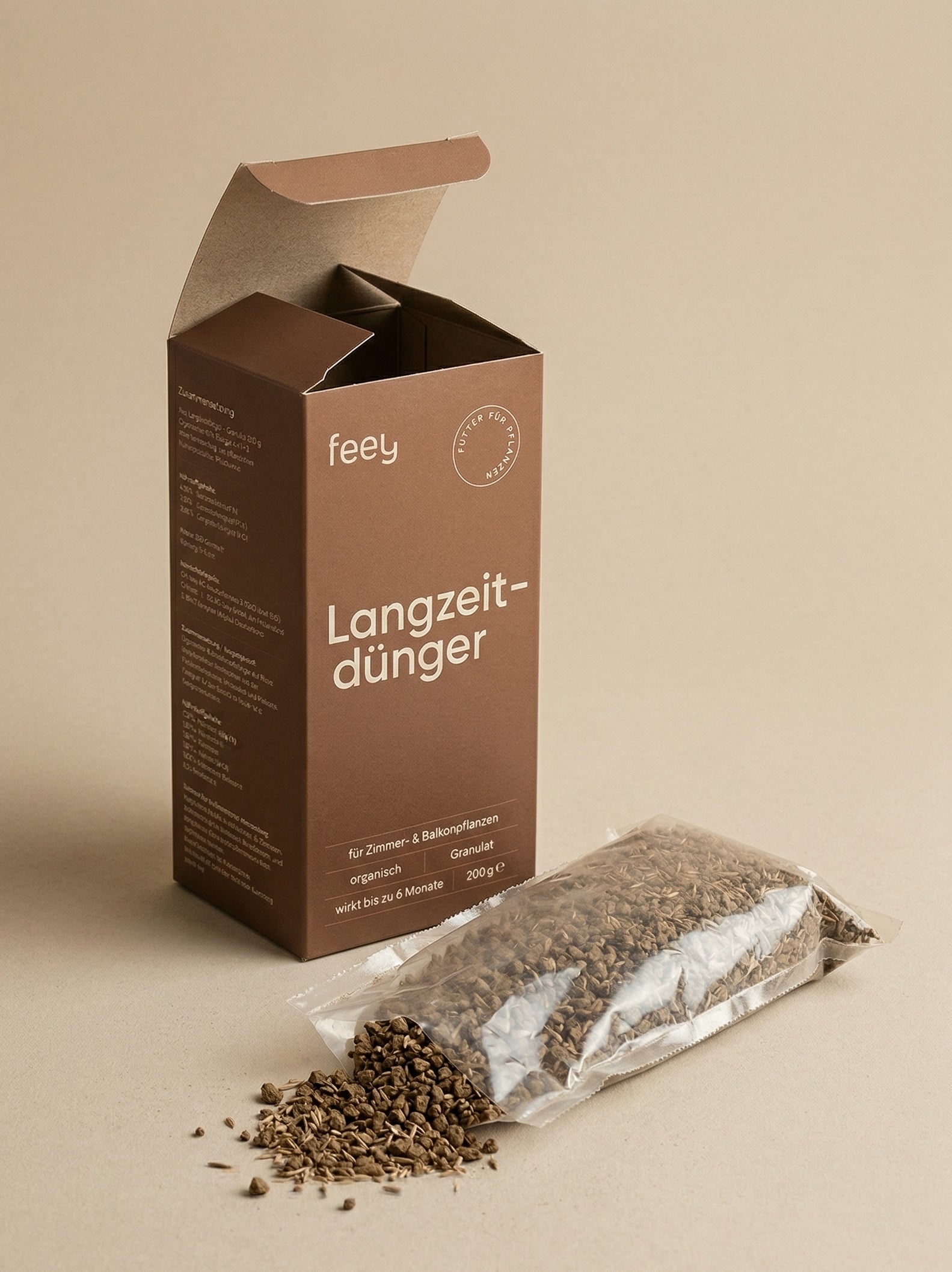 feey Langzeitdünger-Karton mit geöffnetem Deckel und Granulatbeutel daneben, einzelne Körner auf beigem Untergrund verstreut
