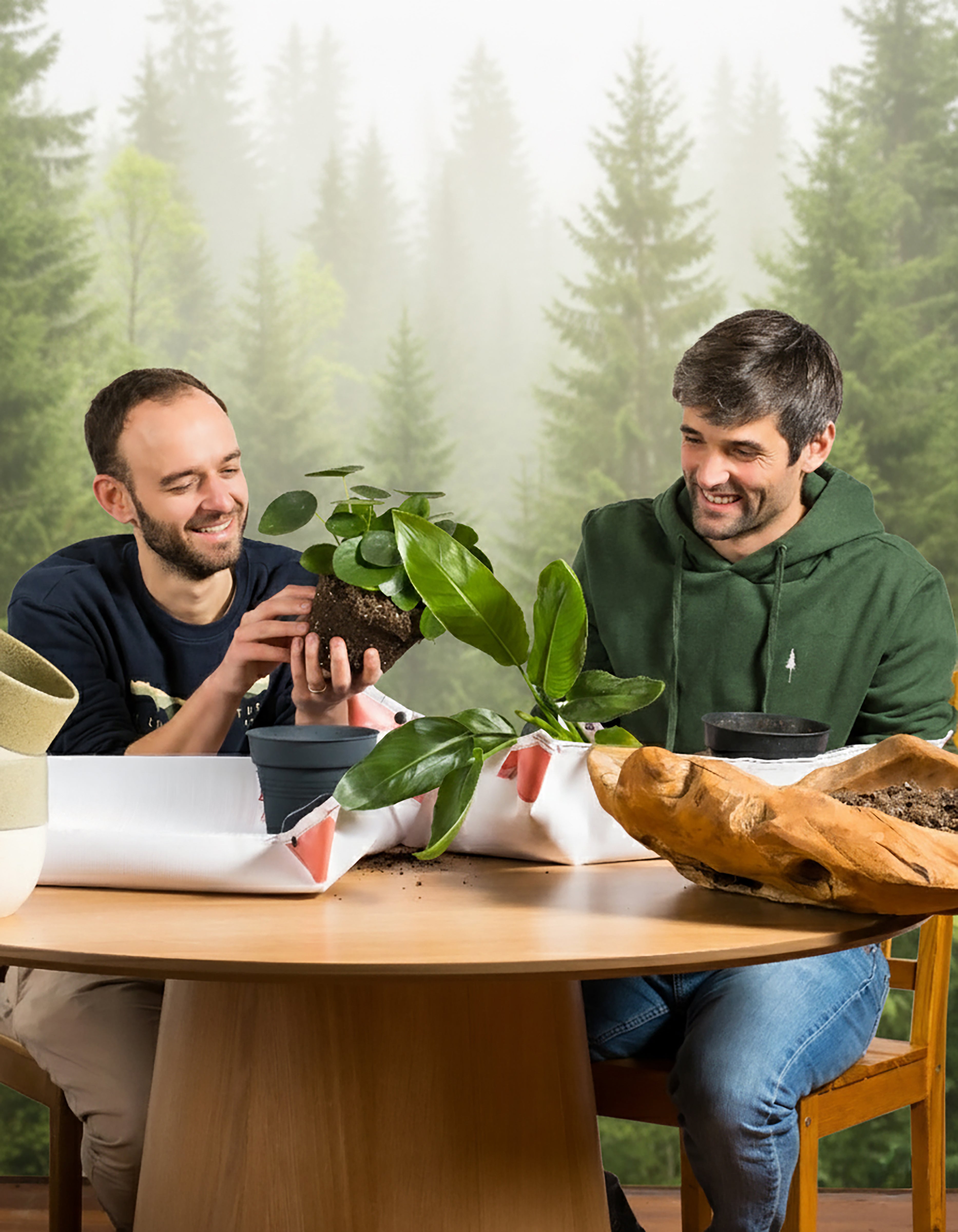 Zwei lächelnde Männer sitzen an einem Holztisch beim Umtopfen von Pflanzen aus dem Pflanzen Adventskalender | NIKIN-Special (Dschungel-Version). Blumentöpfe, Erde und Werkzeuge stehen auf dem Tisch, im Hintergrund sorgt ein Waldwandbild für eine fröhliche Atmosphäre.