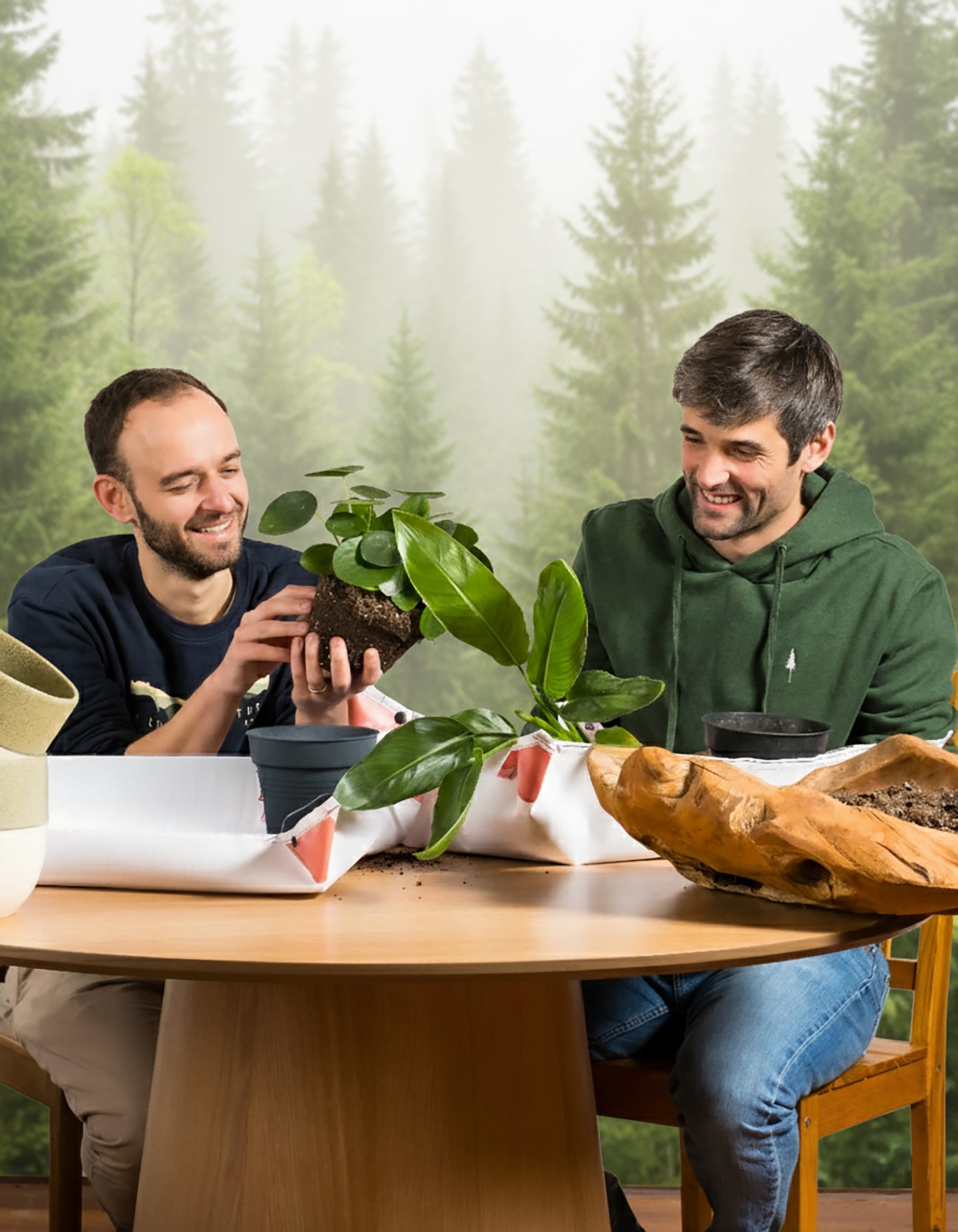 Zwei lächelnde Männer sitzen an einem Holztisch beim Umtopfen von Pflanzen aus dem Pflanzen Adventskalender | NIKIN-Special (Dschungel-Version). Blumentöpfe, Erde und Werkzeuge stehen auf dem Tisch, im Hintergrund sorgt ein Waldwandbild für eine fröhliche Atmosphäre.