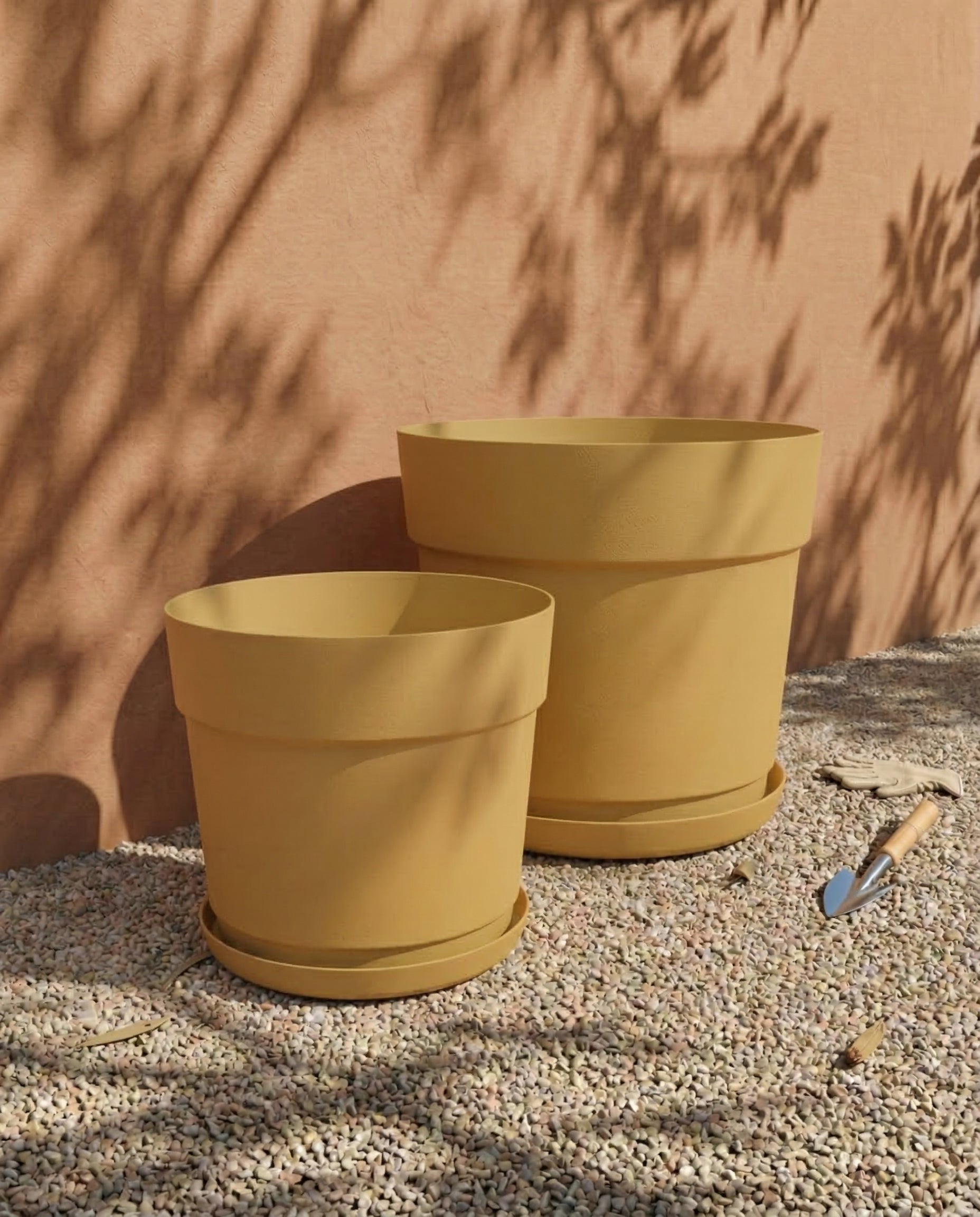 Zwei große, leere, gelbe Pflanzkübel-Sets 'Bloom' (38, 29) aus recyceltem Kunststoff sitzen auf Kies an einer Pfirsichwand, in deren Nähe sich Gartengeräte und heruntergefallenes Laub befinden. Die Schatten von Baumzweigen fallen auf die Mauer und den Boden.