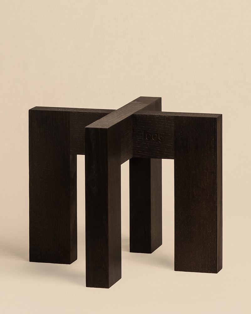 Der minimalistische Pflanzenständer (Heviton | 25 cm Ø) besteht aus zwei ineinandergreifenden dunkelbraunen L-förmigen Holzteilen, die ein abstraktes und symmetrisches Design erzeugen, das an die Ästhetik moderner Holzhocker erinnert. Ein schlichter beiger Hintergrund unterstreicht seine Eleganz und Schlichtheit.