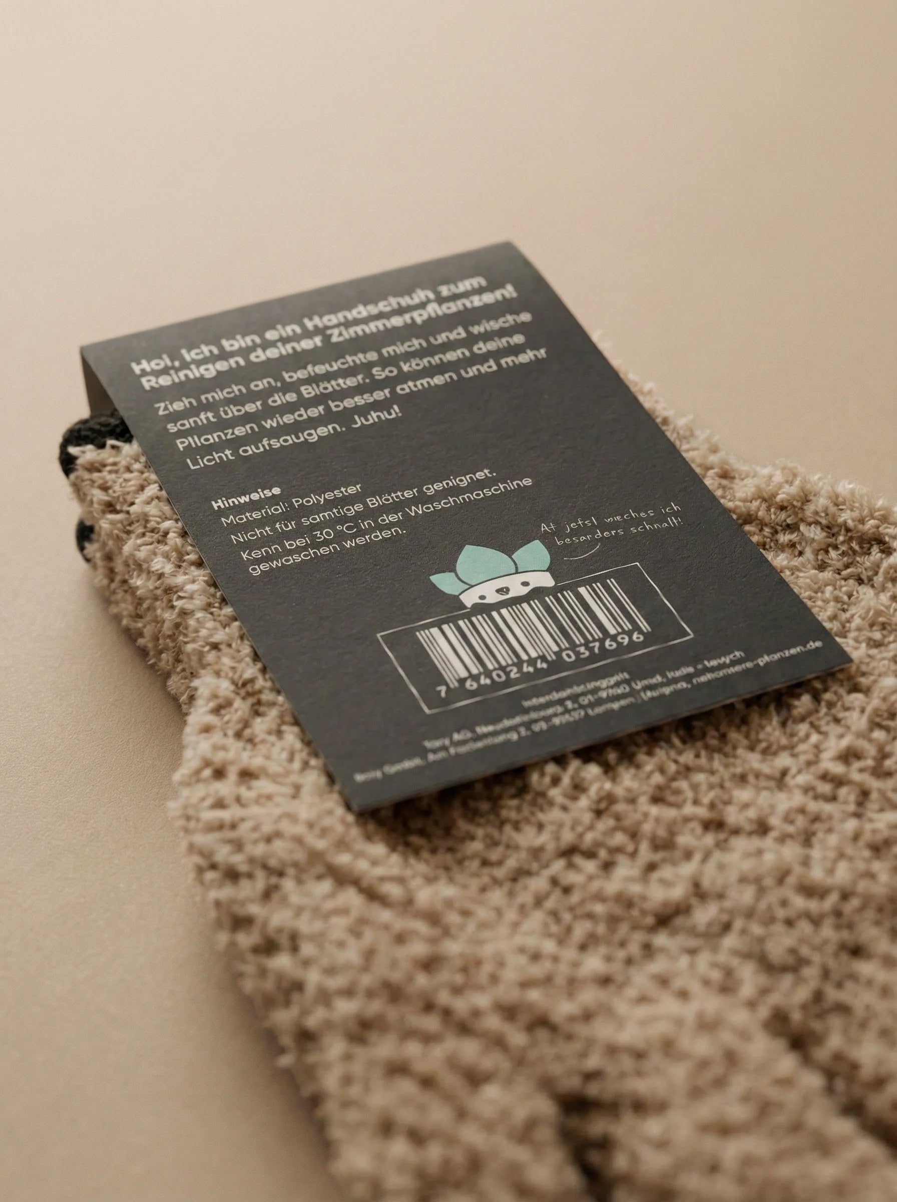 Nahaufnahme eines braunen Staubhandschuhs mit einem schwarzen Etikett in deutscher Sprache, das Pflegehinweise und eine Zeichentrickfigur zeigt. Der Handschuh ist für die Entfernung von Blattstaub bestimmt und liegt auf einer beigen Fläche mit einem Strichcode am unteren Rand des Etiketts.