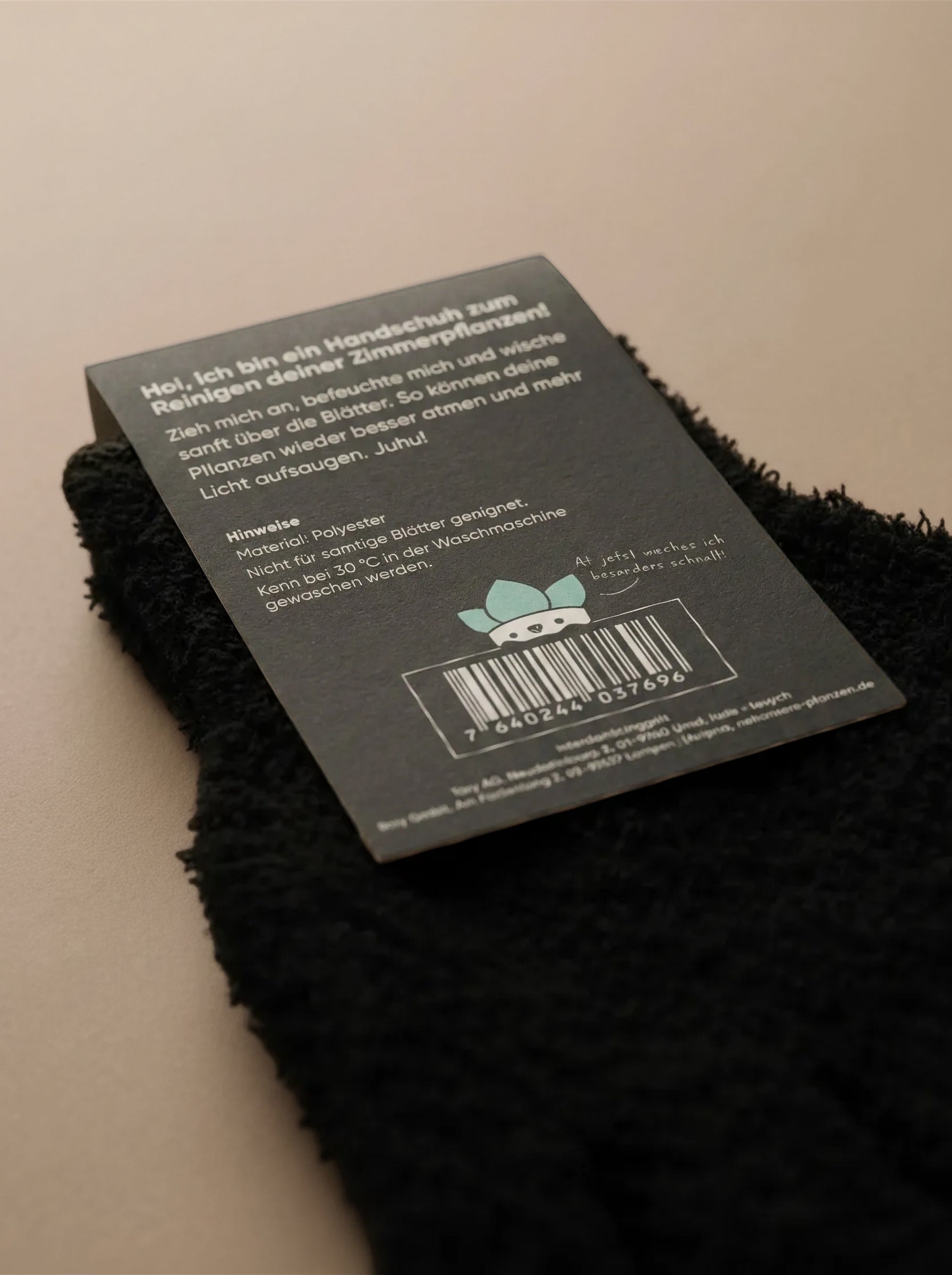 Ein schwarzer Staubhandschuh für Zimmerpflanzen Abstauben ruht auf einer beigen Oberfläche und zeigt ein deutsches Etikett mit Barcode, Anweisungen und einer blauen Tierillustration. Perfekt zum Entfernen von Blattstaub.