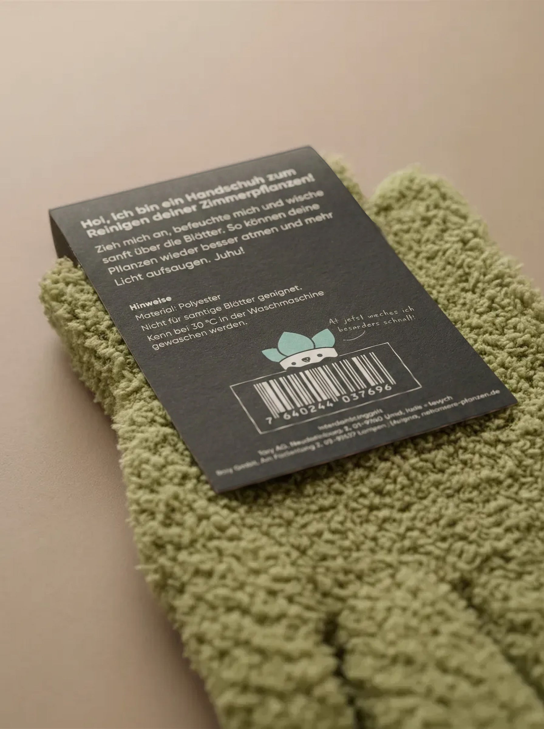 Ein Paar grüner, strukturierter Staubhandschuhe liegt auf einer beigen Fläche. Daran befestigt ist ein schwarzes Etikett mit deutschem Text, einem Strichcode und einer Zeichentrickfigur. Ideal zum sanften Abstauben von Zimmerpflanzen.