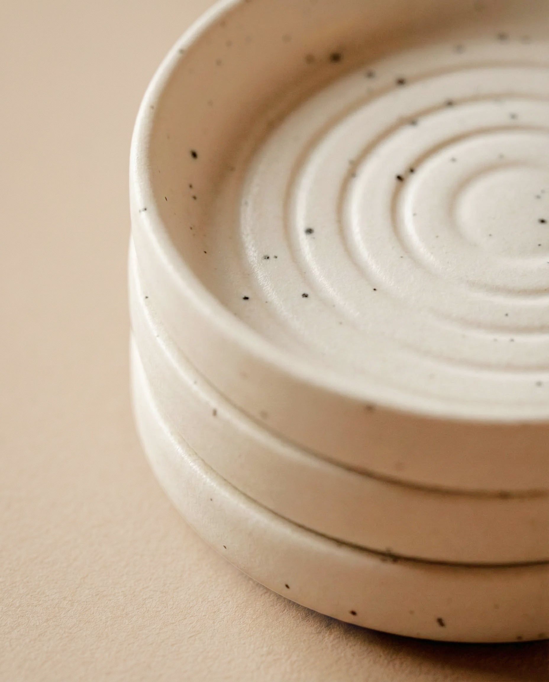 Drei gestapelte Babypflanzen-Untersetzer aus Keramik (Plattino | 3 Stk.) mit einer cremefarbenen, gesprenkelten Oberfläche und konzentrischen Kreismustern sind auf einem neutralen beigen Hintergrund angeordnet.