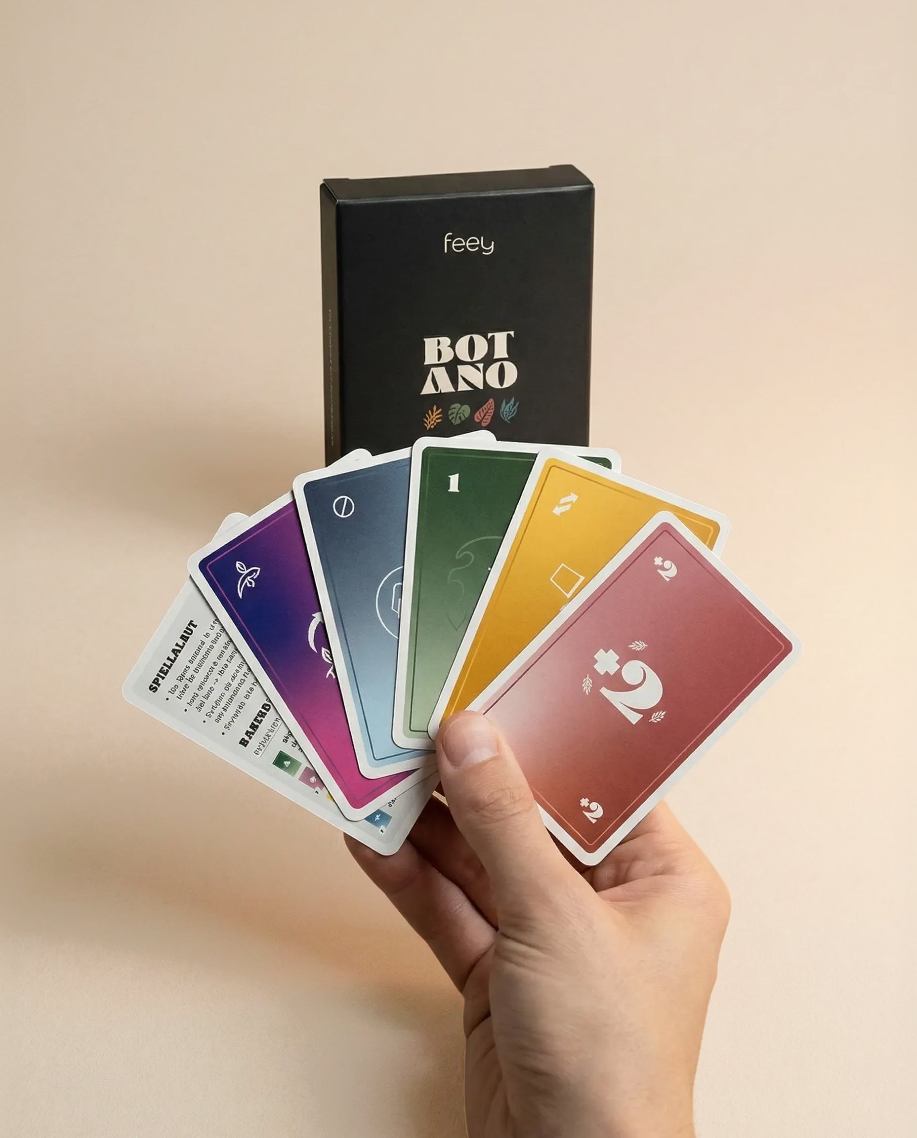 Ein grüner Daumen hält einen Fächer mit bunten, nummerierten Karten vor einer schwarzen Kartenspiel 'Botano'-Box von feey, auf der verschiedene Symbole vor einem neutralen Hintergrund abgebildet sind.