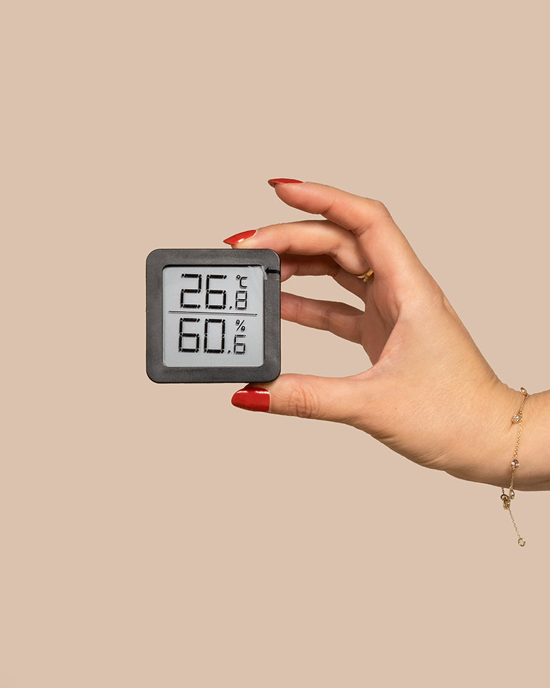 Eine Hand mit rotem Nagellack hält ein kleines Hygrometer, das eine Temperatur von 26,8°C und eine Luftfeuchtigkeit von 60,6% anzeigt. Der Hintergrund ist schlicht beige. Die Hand trägt außerdem ein zartes goldenes Armband am Handgelenk.