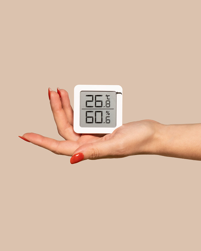 Eine Hand mit roten Nägeln hält ein kleines weißes Hygrometer, das eine Luftfeuchtigkeit von 60,6 % und eine Temperatur von 26,8 °C anzeigt. Der Hintergrund ist beige.