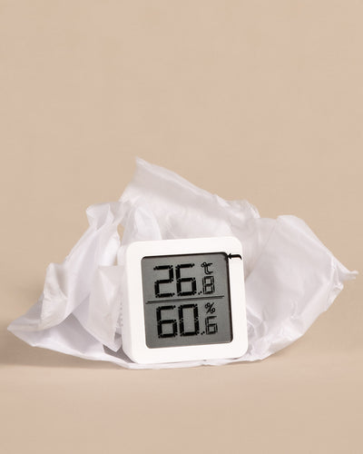 Ein Hygrometer mit einer Temperatur von 26,1 °C und einer Luftfeuchtigkeit von 60,1 % ist auf einem weißen, zerknüllten Seidenpapier und einem beigen Hintergrund abgebildet. Das Gerät ist weiß und hat ein klares Display, auf dem die Messwerte in schwarzen Ziffern angezeigt werden.
