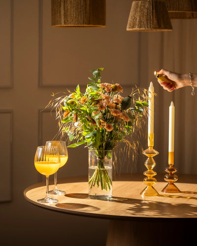 Der Blumenstrauß "Goldene Stunde" in einer Glasvase steht auf einem Holztisch mit Orangensaft und Kerzen. Eine Hand zündet eine Kerze an. Warme Farben und gewebte Lampenschirme sorgen für gemütliche Stimmung.