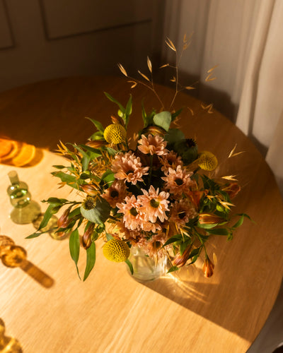 Der Blumenstrauß "Goldene Stunde" aus pinken Gänseblümchen und gelben Billy Balls mit grünen Akzenten steht in einer Glasvase auf einem Holztisch und leuchtet im warmen Licht der goldenen Stunde.