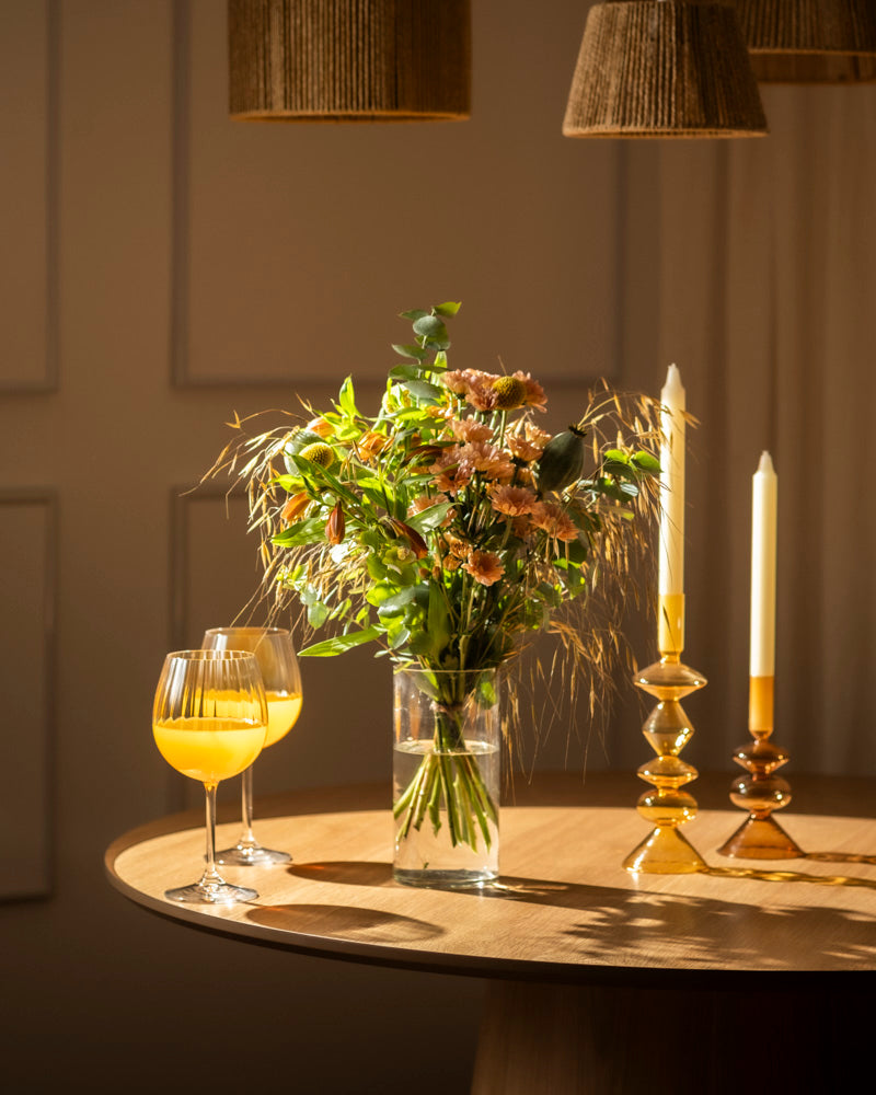 Ein runder Holztisch mit zwei Gläsern Orangensaft, einem Blumenstrauß und zwei Kerzen in Glasbehältern, stimmungsvoll beleuchtet von der warmen Goldenen Stunde-Lampe und sanften Lampenschirmen darüber.