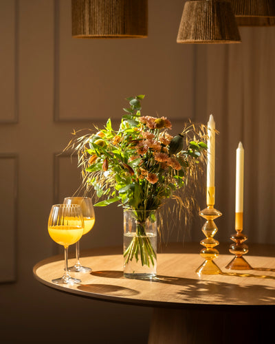 Ein runder Holztisch mit zwei Gläsern Orangensaft, einem Blumenstrauß und zwei Kerzen in Glasbehältern, stimmungsvoll beleuchtet von der warmen Goldenen Stunde-Lampe und sanften Lampenschirmen darüber.