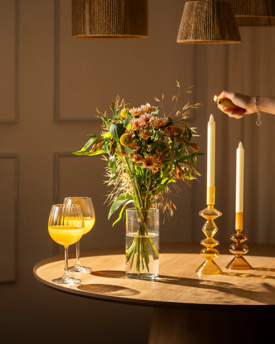 Auf einem runden Holztisch mit warmen Farben stehen Goldene Stunde-Kerzen in goldenen Haltern, eine Vase mit Blumen, zwei Orangensaftgläser und eine Hand, die eine Kerze anzündet, während weiche Schatten geworfen werden.