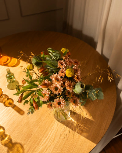 Die Goldene Stunde: Eine kleine Glasvase mit rosafarbenen Gänseblümchen, gelben Bällen, Grünzeug und getrocknetem Gras steht auf einem runden Holztisch in warmen Farbtönen, der in der Nähe eines Vorhangs im Sonnenlicht der goldenen Stunde leuchtet.