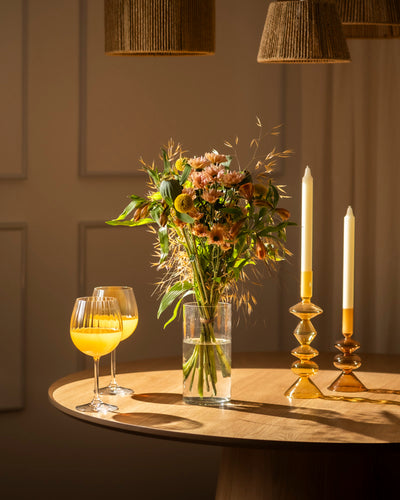 Der runde Holztisch der Goldenen Stunde ist mit zwei Gläsern Orangensaft, einem Blumenstrauß und hohen Kerzen in bernsteinfarbenen Haltern gedeckt, die unter Hängelampen warm leuchten.
