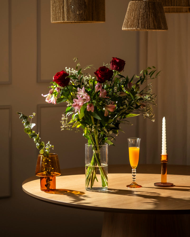 Ein prächtiger Blumenstrauß "Ich liebe Dich" mit roten Rosen auf einem runden Holztisch - begleitet von Orangensaft, weißer Kerze im bernsteinfarbenen Halter und Bernsteinvase - ein perfektes Geschenk, um Liebe auszudrücken.