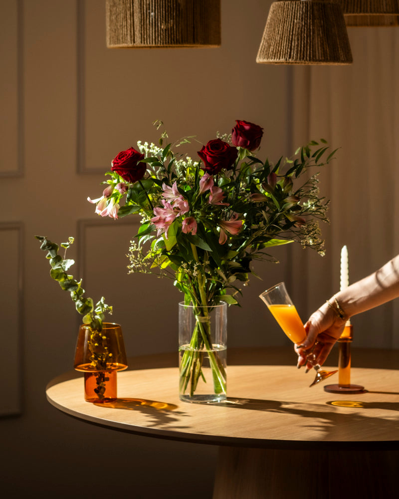 Der "Ich liebe Dich"-Blumenstrauß aus roten und rosafarbenen Blumen steht auf einem runden Holztisch mit Braunglasvasen und einer Kerze, während eine Hand mit einem orangefarbenen Getränk zur gemütlichen, romantischen Stimmung beiträgt.