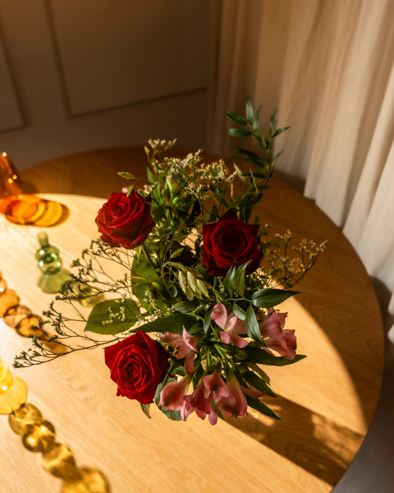 Ein wunderschöner "Ich liebe Dich"-Blumenstrauß aus roten Rosen und rosafarbenen Blumen mit Grünzeug liegt auf einem runden Holztisch, der in Sonnenlicht getaucht ist - ein ideales Geschenk, um Ihre Liebe auszudrücken, mit leuchtenden Glasvasen im Hintergrund.