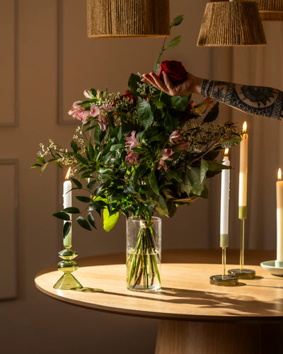 Ein tätowierter Arm stellt eine rote Rose in eine hohe Vase mit rosa Blumen und Grünzeug mit der Aufschrift "Ich liebe Dich" auf einen runden Holztisch, neben drei brennende Kerzen in grünen Haltern - das perfekte Geschenk, um "Ich liebe Dich" zu sagen.