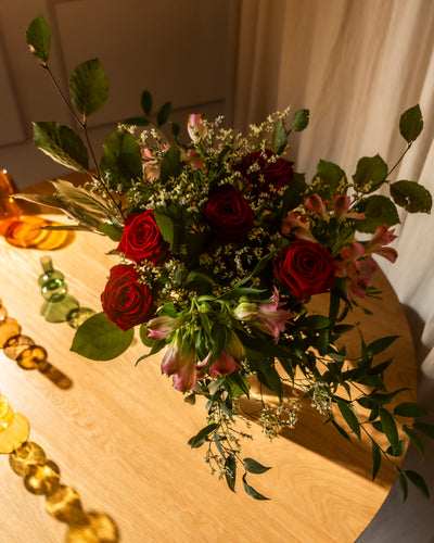 Der "Ich liebe Dich"-Blumenstrauß - rote Rosen, rosa Lilien und Grünzeug - steht in warmem Licht auf einem Holztisch mit gelbem und grünem Glasdekor daneben und ist das perfekte Geschenk, um "Ich liebe dich" zu sagen.