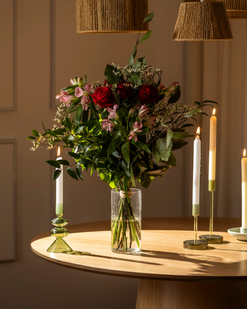 Der Blumenstrauß "Ich liebe Dich" zeigt rote und rosafarbene Blumen mit grünem Laub in einer durchsichtigen Vase, die auf einem Holztisch mit beleuchteten Kerzenständern steht - warm beleuchtet für das perfekte romantische Geschenk.