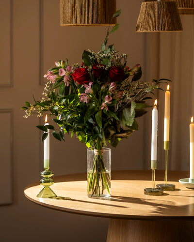 Der Blumenstrauß "Ich liebe Dich" zeigt rote und rosafarbene Blumen mit grünem Laub in einer durchsichtigen Vase, die auf einem Holztisch mit beleuchteten Kerzenständern steht - warm beleuchtet für das perfekte romantische Geschenk.