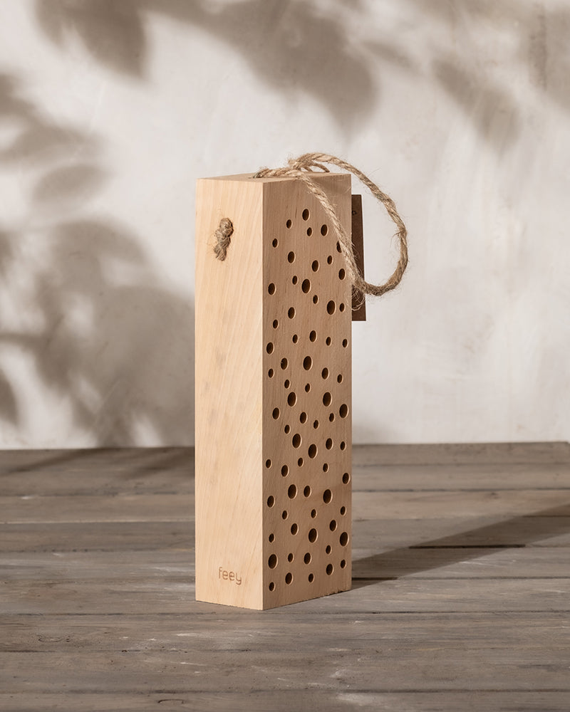 Das Insektenhotel „Erbienbi“ mit seinem minimalistischen rechteckigen Holzdesign mit kleinen Löchern und einem Seilgriff ruht elegant auf einer Holzoberfläche. Das Spiel aus geflecktem Sonnenlicht und weichen Schatten verstärkt seine natürliche Ästhetik noch weiter.