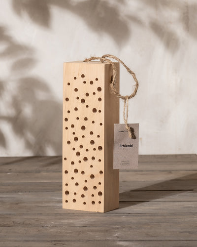 Auf einer Holzoberfläche steht das Insektenhotel „Erbienbi“, ein Holzblock mit zahlreichen Löchern, der als perfekter Unterschlupf für Wildbienen dient. An der Oberseite befindet sich eine Seilschlaufe mit einem kleinen Schild, während Schatten von nahe gelegenen Objekten dem strukturierten Hintergrund Tiefe verleihen.