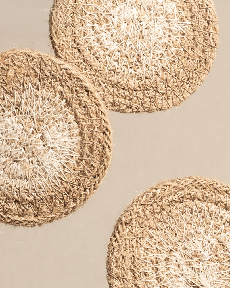Drei runde Babypflanzen-Untersetzer aus Jute, handgewebt aus Naturfasern, sind diagonal auf einem neutralen Hintergrund angeordnet. Ihr strukturiertes Design besteht aus hell- und mittelbraunen Fäden in einem konzentrischen Muster, ideal für die Präsentation von Babypflanzen.