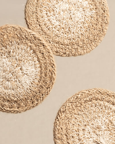 Drei runde Babypflanzen-Untersetzer aus Jute, handgewebt aus Naturfasern, sind diagonal auf einem neutralen Hintergrund angeordnet. Ihr strukturiertes Design besteht aus hell- und mittelbraunen Fäden in einem konzentrischen Muster, ideal für die Präsentation von Babypflanzen.