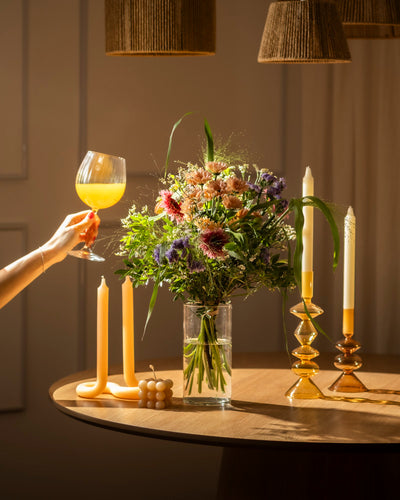 Eine Hand hält ein großes Glas mit einem gelb-orangenen Getränk neben einem runden Tisch mit dem Strauß "So kunterbunt wie Du" in einer Vase, hohen Kerzenhaltern und dekorativen Kerzen, die alle warm beleuchtet sind.
