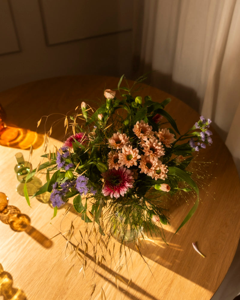 Der Strauß "So kunterbunt wie Du", gefüllt mit rosa, violetten und magentafarbenen Blumen sowie grünem Laub, steht auf einem runden Holztisch im warmen Sonnenlicht, das sanfte Schatten wirft und eine gemütliche Atmosphäre schafft.