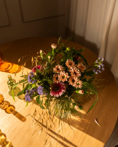 Der Strauß "So kunterbunt wie Du", gefüllt mit rosa, violetten und magentafarbenen Blumen sowie grünem Laub, steht auf einem runden Holztisch im warmen Sonnenlicht, das sanfte Schatten wirft und eine gemütliche Atmosphäre schafft.