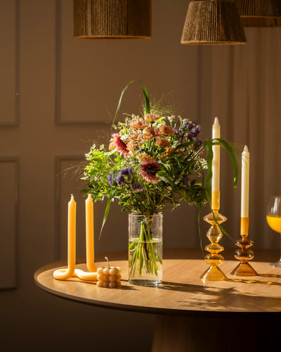 Der bunte Blumenstrauß "So kunterbunt wie Du" steht in einer klaren Glasvase auf einem runden Holztisch mit Kerzen, Kerzenhaltern und einem Glas Orangensaft im warmen, gemütlichen Licht.
