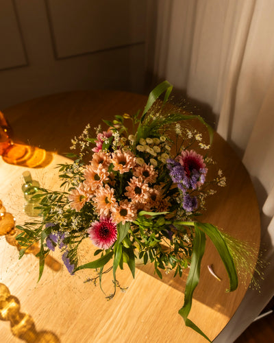 Der bunte Blumenstrauß "So kunterbunt wie Du" mit rosa, lila und weißen Blüten und grünen Blättern steht auf einem runden Holztisch, der durch ein nahes Fenster in warmes Tageslicht getaucht wird.